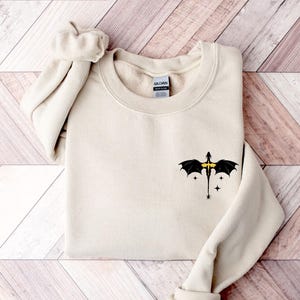 Puede incluir: Una sudadera de color crema con un gr&aacute;fico de drag&oacute;n negro con detalles amarillos en el pecho izquierdo. El drag&oacute;n est&aacute; volando con las alas extendidas y tiene tres estrellas a su alrededor.