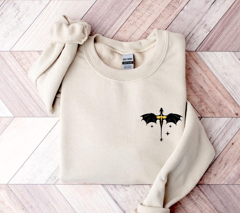 Puede incluir: Una sudadera de color crema con un gr&aacute;fico de drag&oacute;n negro con detalles amarillos en el pecho izquierdo. El drag&oacute;n est&aacute; volando con las alas extendidas y tiene tres estrellas a su alrededor.