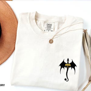 May include: Ivory t-shirt with a black dragon graphic and a gold necklace. The shirt has a crew neck and short sleeves. The text "COMFORT COLORS" is on the tag.