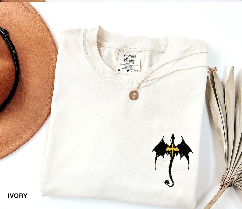 May include: Ivory t-shirt with a black dragon graphic and a gold necklace. The shirt has a crew neck and short sleeves. The text "COMFORT COLORS" is on the tag.