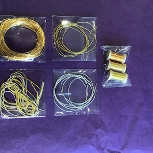 May include: Five clear plastic bags containing spools of gold and silver wire. The wire is in various thicknesses and is arranged in a variety of ways. The bags are on a purple surface.