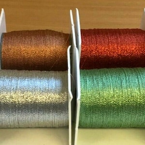 May include: Eight spools of metallic embroidery thread in various colours, including gold, silver, green, and copper. The thread is wound around white spools and is displayed on a wooden surface.