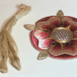 May include: A close-up of a hand-stitched embroidered rose patch with a gold centre and pink and white petals. A skein of beige embroidery floss is also pictured.