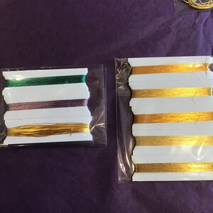 May include: Two packages of embroidery floss, one with green, purple, and gold floss, and the other with gold floss.
