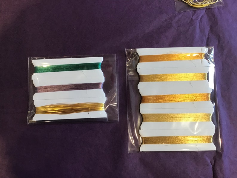 May include: Two packages of embroidery floss, one with green, purple, and gold floss, and the other with gold floss.