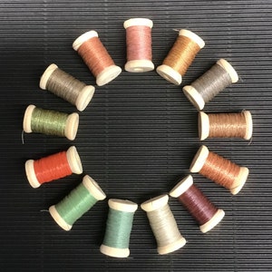 May include: A collection of 12 spools of thread in various colours, including brown, pink, green, and silver. The spools are arranged in a circle on a black background.