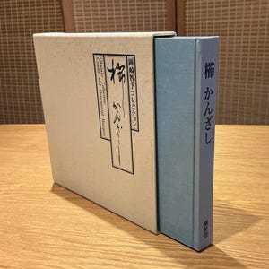 May include: A book and slipcase set. The slipcase is off-white with Japanese text and a black and white illustration. The book is light blue with Japanese text. The set is on a wooden surface.