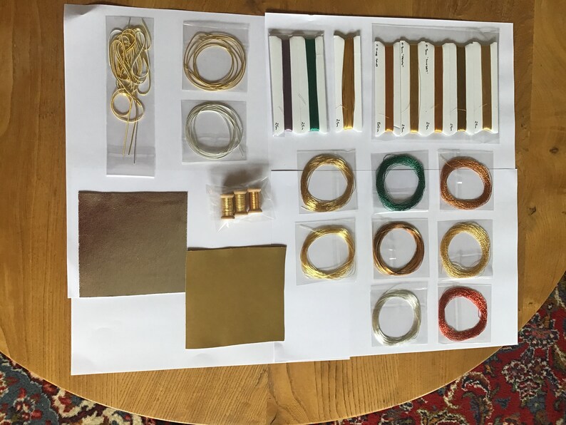 May include: Assortment of embroidery floss, thread, and needles in various colors, including gold, green, and copper. There are also two pieces of gold-colored fabric.