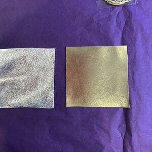 May include: Two square pieces of fabric, one silver and one gold, on a purple background.