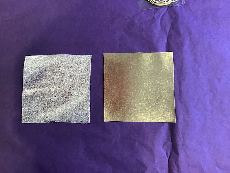 May include: Two square pieces of fabric, one silver and one gold, on a purple background.