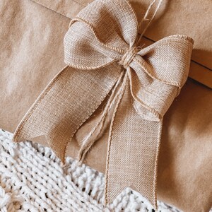 Könnte beinhalten: Nahaufnahme eines Geschenks, das in braunes Papier verpackt und mit einer Schleife aus Juteband verziert ist. Die Schleife besteht aus einem breiten, strukturierten Band und Bindfaden. Das Geschenk liegt auf einer weißen, gestrickten Decke.