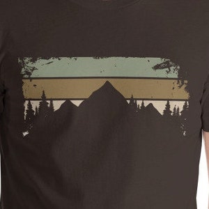 May include: A brown t-shirt featuring a vintage-style mountain range graphic. The graphic depicts a silhouette of mountains with a faded, layered background of green, brown, and white.