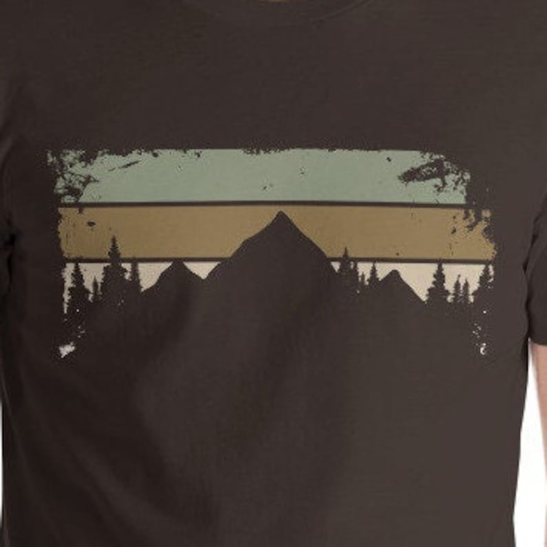 May include: A brown t-shirt featuring a vintage-style mountain range graphic. The graphic depicts a silhouette of mountains with a faded, layered background of green, brown, and white.