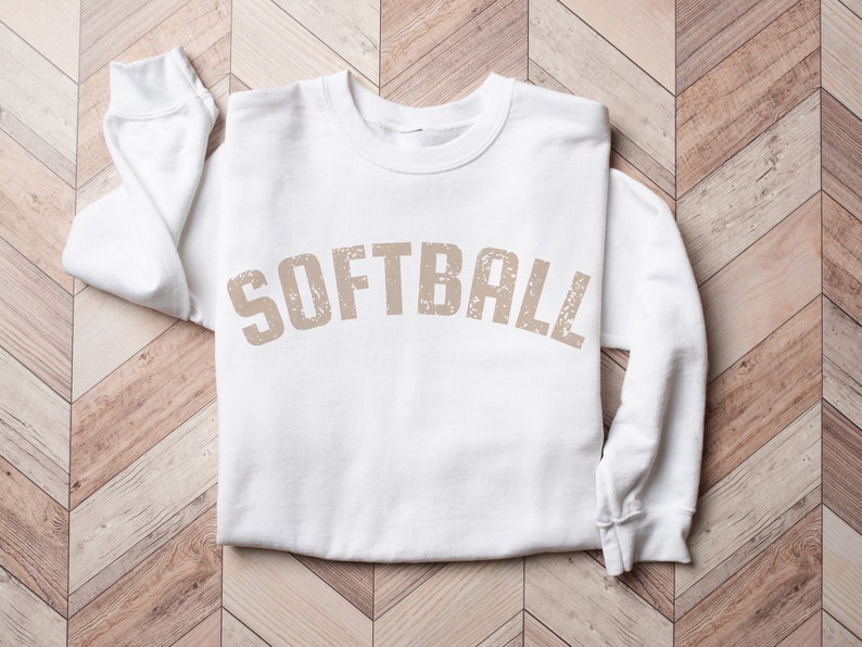 May include: White sweatshirt with the word "SOFTBALL" printed in a distressed brown font.