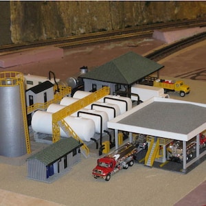 May include: A model industrial complex with a silver silo, white cylindrical tanks, and buildings with green and grey roofs. Yellow ladders and walkways link the structures. Red and yellow trucks are also visible.