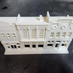 May include: A 3D-printed model of a row of buildings, with detailed architectural features. The off-white model includes windows, doors, and decorative trim. The buildings have a classic, historical appearance.