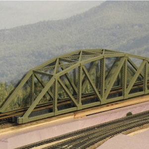 Puede incluir: Un puente de tren modelo de metal verde con un diseño de armadura. El puente está colocado sobre una base de madera y tiene dos juegos de vías.