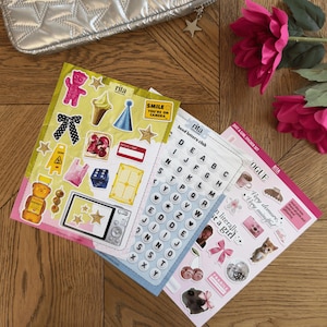 K&ouml;nnte beinhalten: Drei Stickerb&ouml;gen mit verschiedenen Designs, darunter Buchstaben, Essen und S&auml;tze wie "SMILE YOU'RE ON CAMERA". Die Aufkleber sind farbenfroh und zeigen Illustrationen einer Kamera, eines Eis und eines B&auml;ren. Eine silberne Tasche und rosa Blumen sind im Hintergrund.