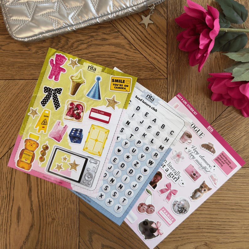 K&ouml;nnte beinhalten: Drei Stickerb&ouml;gen mit verschiedenen Designs, darunter Buchstaben, Essen und S&auml;tze wie "SMILE YOU'RE ON CAMERA". Die Aufkleber sind farbenfroh und zeigen Illustrationen einer Kamera, eines Eis und eines B&auml;ren. Eine silberne Tasche und rosa Blumen sind im Hintergrund.