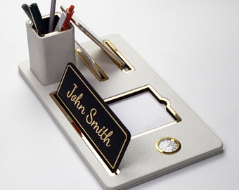 Könnte beinhalten: Weißer Leder-Schreibtischorganizer mit goldener Einfassung. Er verfügt über einen Stiftehalter, einen Handyständer, einen Bilderrahmen und eine kleine Uhr. Der Namensschildhalter trägt die Aufschrift "John Smith".