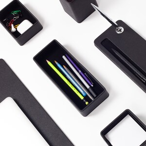 May include: Black leather desk organizer set with compartments for pens, pencils, and other office supplies. The set includes a pen holder, a pencil holder, a small tray, and a larger tray.