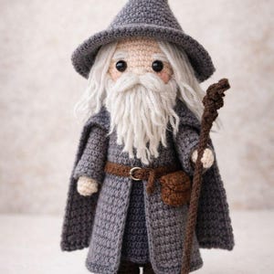 Può includere: Bambola mago all'uncinetto, grigia e marrone. La bambola ha una lunga barba bianca, un cappello grigio e un bastone. Indossa un mantello grigio e una cintura marrone con una piccola borsa. Fatto a mano.