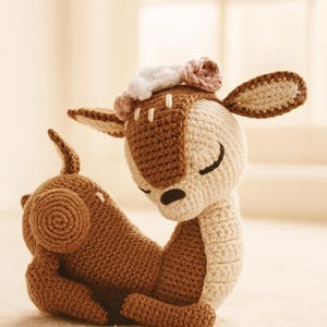 Può includere: Un giocattolo fatto a mano all'uncinetto a forma di cervo, nei toni del marrone e del crema, con un piccolo dettaglio floreale sulla testa. Il cervo è in posizione di riposo, con gli occhi chiusi. Il giocattolo è fatto di filato morbido.