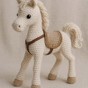 Può includere: Un cavallo giocattolo all'uncinetto color crema con sella e briglia marroni. Il cavallo ha criniera e coda soffici e occhi neri. Il giocattolo è alto circa 25 cm.