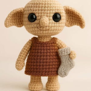 May include: A crocheted doll in the shape of a character with large ears, big black eyes, and a small nose. The doll wears a brown dress and holds a gray sock. The doll is made of beige yarn.