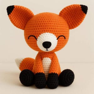 May include: A crocheted fox toy in orange, white, and black. The fox has large ears with black tips, a white chest, and black paws. The fox is sitting with its legs crossed and has a smiling face.