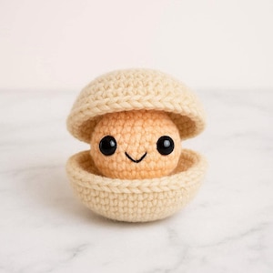 Può includere: Una conchiglia di vongola all'uncinetto beige, con una creatura sorridente color pesca all'interno. La conchiglia è aperta nella parte superiore. La creatura ha occhi neri e un sorriso cucito. Lo sfondo è una superficie di marmo chiaro.