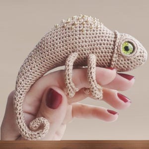 May include: A crocheted chameleon in beige yarn, featuring a beaded spine and vivid green eyes. The handmade reptile has curled feet and a curled tail. A unique, handcrafted item.