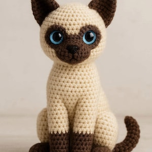 Può includere: Figurina di gatto siamese all'uncinetto con corpo color crema e dettagli marrone scuro su orecchie, muso, zampe e coda. Il gatto ha grandi occhi blu brillante ed è seduto.