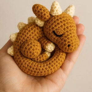 Può includere: Un giocattolo a forma di dinosauro all'uncinetto fatto a mano, nei toni del giallo senape e crema. Il dinosauro amigurumi è arrotolato, con gli occhi chiusi. Il giocattolo è tenuto in mano.