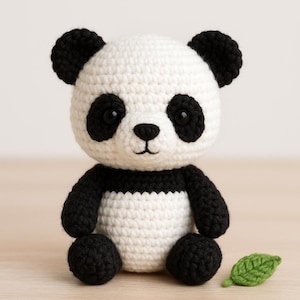 Può includere: Un orsetto panda all'uncinetto, bianco e nero. Il panda ha orecchie, occhi, braccia e gambe nere, con testa e corpo bianchi. Una piccola foglia verde all'uncinetto è vicina. Un giocattolo fatto a mano.