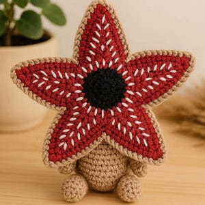 Può includere: Una figura di Demogorgon fatta a mano all'uncinetto. La figura presenta una testa rossa a forma di stella con dettagli bianchi e un centro nero. Il corpo e le gambe sono all'uncinetto in colore beige. La figura è esposta su una superficie di legno.