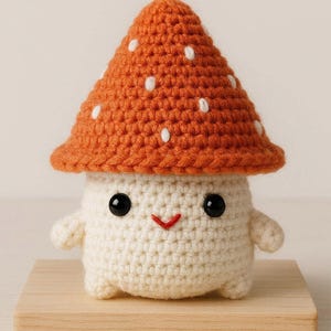Può includere: Una figura di fungo all'uncinetto con un cappello arancione punteggiato di punti bianchi. Il fungo ha un corpo bianco, occhi neri e un sorriso rosso cucito. È esposto su un piccolo blocco di legno.