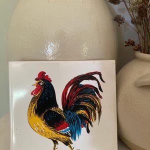 May include: A square ceramic tile featuring a colorful rooster illustration. The rooster has a red comb, yellow body, black and blue feathers, and a vibrant red and blue tail. The tile is signed "R. Howard".