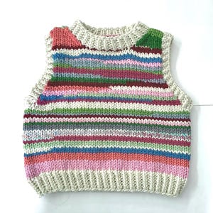 Può includere: Un gilet lavorato a maglia a righe multicolore, con righe rosa, blu, verdi e grigie. Il gilet ha un orlo e un colletto a coste. Le righe sono orizzontali. Il capo è senza maniche.