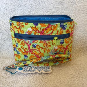 May include: A small, rectangular pouch with a wrist strap. The pouch features a vibrant green fabric with a colorful floral pattern. It has two zippered compartments with blue trim and a blue interior. The wrist strap has the word "LOVE" on it.