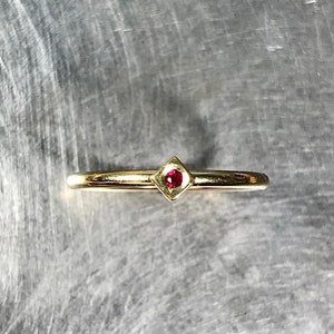 May include: A delicate gold band ring with a small, square-shaped ruby gemstone set in the center.