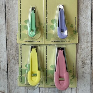 May include: Four Clover fusible bias tape makers in green, purple, yellow, and pink. Each tool is packaged on a card with instructions and the Clover logo. The tools are designed to create fusible bias tapes.