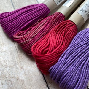 May include: Three spools of embroidery floss in shades of purple, red, and pink. The spools are wrapped in brown paper with Japanese text and barcodes.