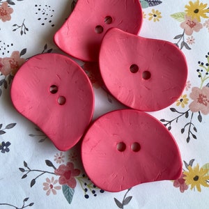 May include: Four pink, two-hole, petal-shaped buttons. The buttons are made of a textured material and have a slightly raised edge.
