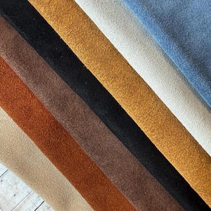 May include: A collection of suede fabric swatches in various colors, including beige, rust, brown, black, tan, gold, white, and blue. The swatches are arranged diagonally, showcasing the texture and color variations.