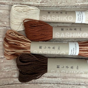 May include: Four spools of Sashiko thread in various earth-tone colors: cream, rust, light brown, and dark brown. Each spool is labeled with Japanese text and the word "SASHIKO". The thread is 100% cotton and made in Japan.