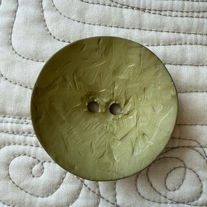 May include: A large, round, olive-green button with two holes. The button has a textured surface with visible scratches. The button is set against a light-colored, quilted fabric background.