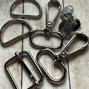 May include: A collection of silver-toned metal hardware components including two D-rings, two swivel lobster claw clasps with oval loops, one rectangular slider buckle, and a small cluster of circular magnetic snap fasteners with metal backing plates.