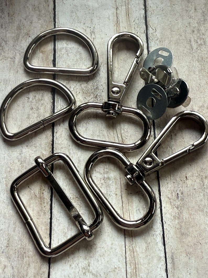 May include: A collection of silver-toned metal hardware components including two D-rings, two swivel lobster claw clasps with oval loops, one rectangular slider buckle, and a small cluster of circular magnetic snap fasteners with metal backing plates.