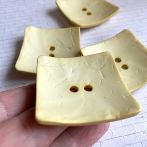 May include: Four square, pale yellow buttons with a slightly concave surface and a textured, crackled finish. Each button features two centered holes for attachment, arranged horizontally in the middle of the square shape.